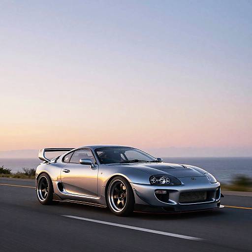 Photograph of a silver, high-performance sports car with a rear spoiler driving on a coastal road during sunset.