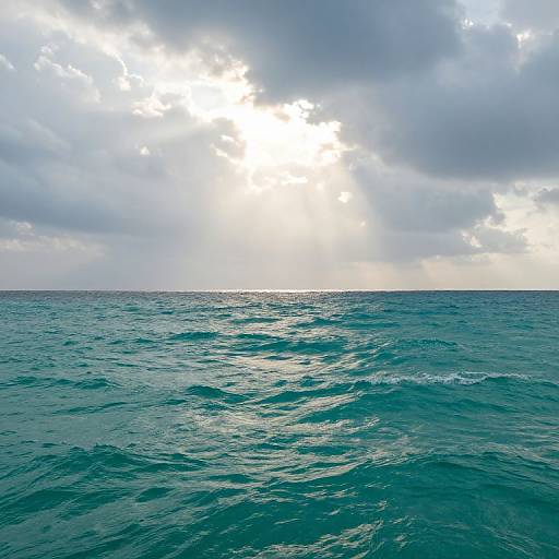 Photograph of a vast, turquoise ocean with gentle waves under a cloudy sky, sunlight breaking through, illuminating the horizon.
