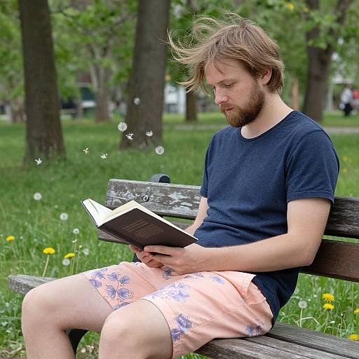 Photograph of a bearded man with light brown hair, wearing a navy t-shirt and peach shorts, reading a book on a park bench, surrounded