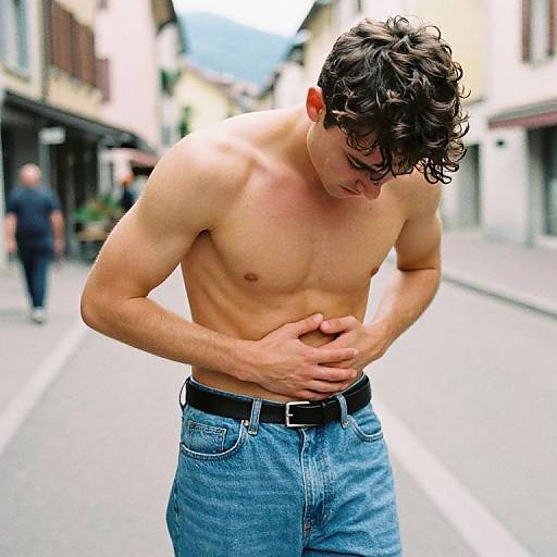 Photograph of a shirtless, muscular young man with curly brown hair, wearing blue jeans and a black belt, standing on a sunny, narrow street