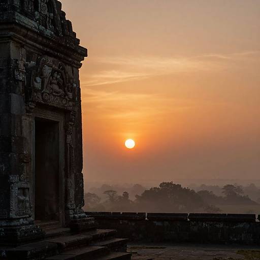 Photograph of a stone temple at sunset, with an orange and yellow sky, a bright sun, and misty distant trees. The temple's intricate