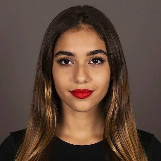 Photograph of a young woman with long, straight brown hair with blonde highlights, wearing red lipstick and black top, against a gray background.