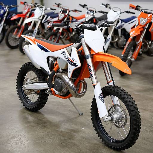 Photograph of a white and orange off-road motorcycle with black tires, surrounded by similar motorcycles in a warehouse.