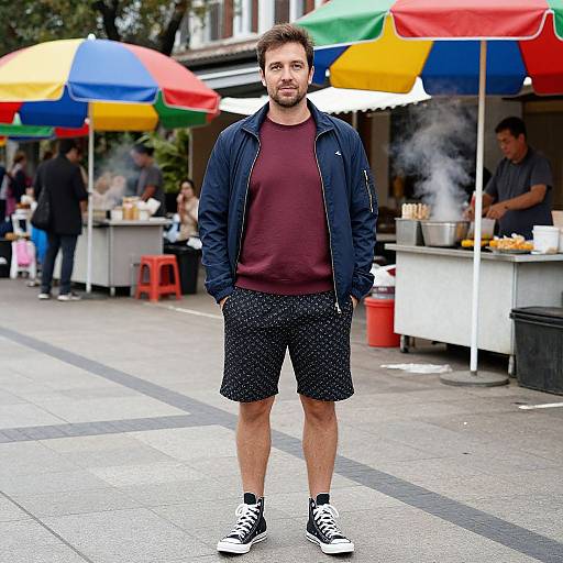 Photograph of a bearded man in a maroon shirt, black jacket, and polka-dot shorts, standing in front of colorful food stalls with