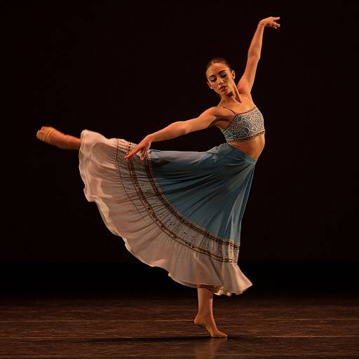Photograph of a ballerina performing a graceful arabesque on stage, wearing a blue and white striped crop top and flowing skirt, against a