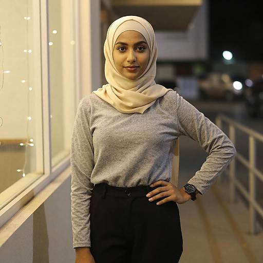 Photograph of a young woman with light brown skin, wearing a beige hijab, grey long-sleeve top, and black pants, standing outdoors
