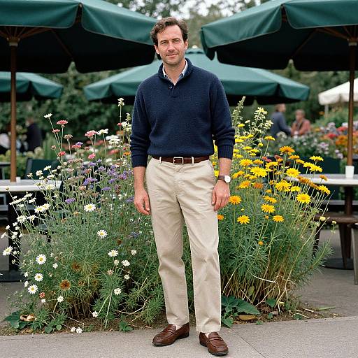 Photograph of a smiling man in a navy sweater, beige pants, and brown shoes, standing amidst colorful flowerbeds under green umbrellas.