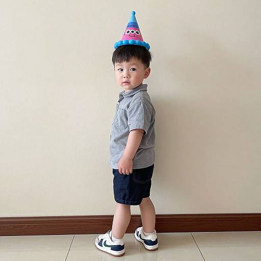 Photograph of a young Asian boy in a gray shirt, black shorts, white sneakers, blue and pink party hat, standing against a beige wall.