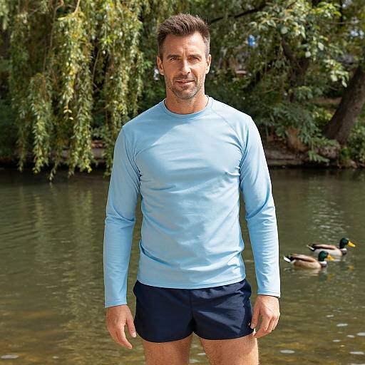 Photograph of a muscular, fair-skinned man with short brown hair, wearing a light blue long-sleeve shirt and navy shorts, standing in