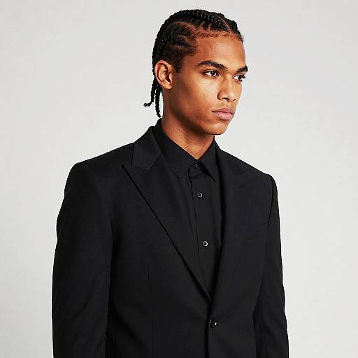 Photograph of a young Black man with braided hair, wearing a black suit and black shirt, looking to the right against a white background.