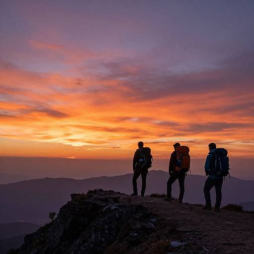 Photograph of three silhouetted hikers with backpacks standing on a rocky ridge at sunset, featuring a vibrant orange and purple sky.