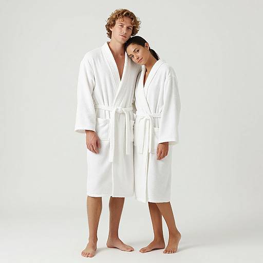 Photograph of a tall, curly-haired man and a shorter woman, both wearing white bathrobes, standing barefoot against a plain white background.