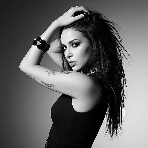Black-and-white photograph of a tattooed, gothic woman with long, messy hair, wearing a black tank top and wrist bracelet, posing with arms