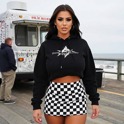 Photograph of a curvy woman with dark hair, wearing a black hoodie and black-and-white checkered skirt, standing in front of a food truck