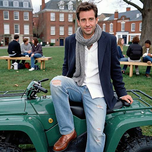 Photograph of a casually dressed man with short brown hair, wearing a black blazer, white shirt, gray scarf, and ripped jeans, sitting on