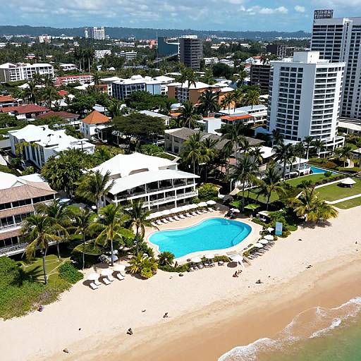 Aerial photograph of a sunny coastal cityscape featuring a white-roofed resort with a blue swimming pool, surrounded by palm trees, white sand beach