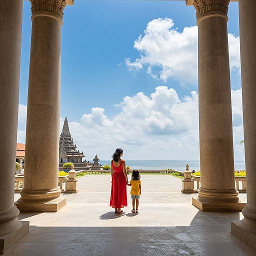 Photograph of a woman in a red dress and a child in a yellow dress, walking through ancient stone pillars towards a coastal temple under a bright blue