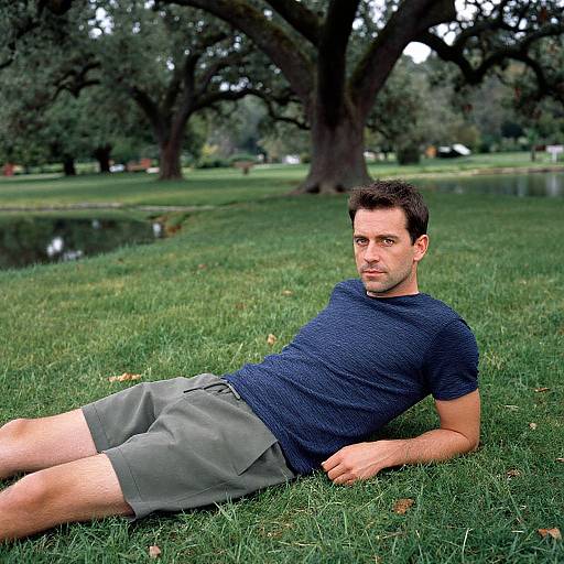 Photograph of a handsome, dark-haired man in a navy t-shirt and gray shorts, reclining on grass in a park with large trees and a