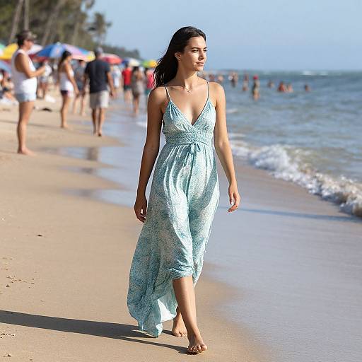 Photograph of a young woman with long dark hair walking on a sunny beach in a light blue, patterned sundress, with people and colorful umb