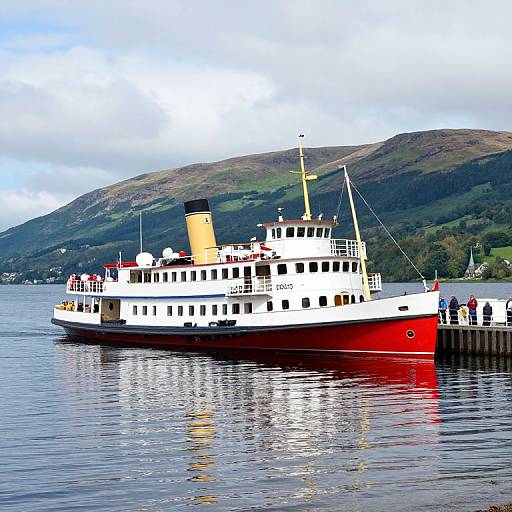 Photograph of a red and white ferry docked on a calm lake, with a mountainous green landscape in the background.