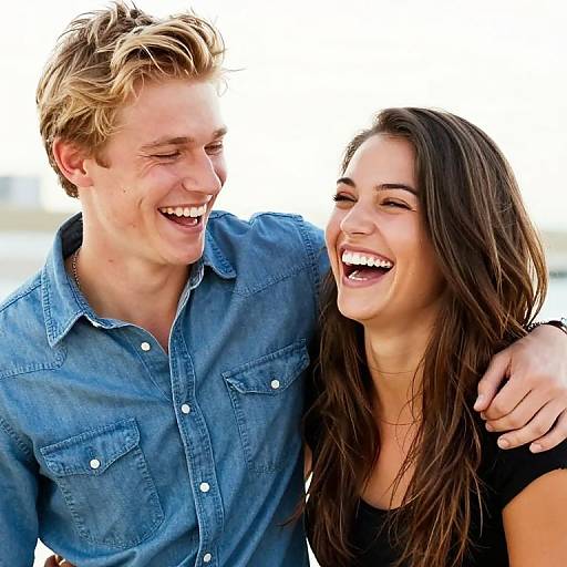 Photograph of a smiling couple; blond man in blue denim shirt, brown-haired woman in black top, standing close, arm around each other. Bright