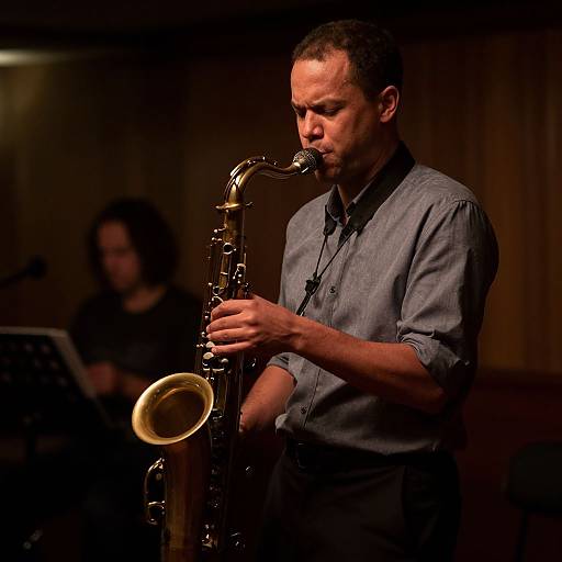 Photograph of a focused male saxophonist with short hair, wearing a gray button-up shirt, playing in a dimly lit room. Blurred