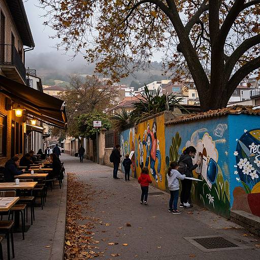 Photograph of a colorful, graffiti-covered alleyway with people walking, autumn leaves, outdoor cafe tables, and trees; misty, cloudy sky in