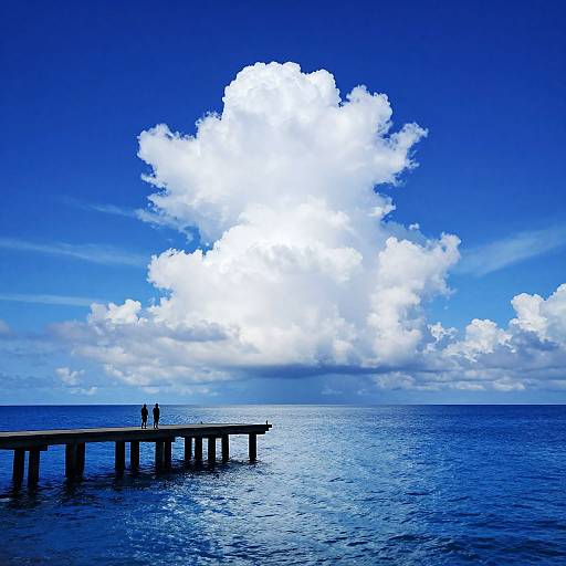 Photograph of a wooden pier extending into a vast blue ocean, with two small figures standing on the pier, under a large, bright white cumulus