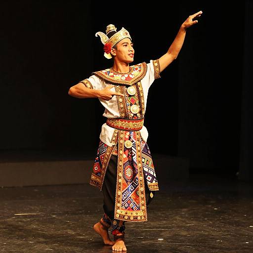 Photograph of a dancer in colorful traditional tribal attire, barefoot, pointing upwards, standing on a dark stage with a black background.