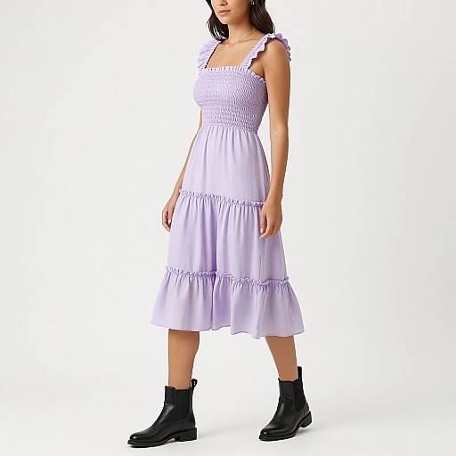 Photograph of a woman in a white, sleeveless, smocked dress with ruffle trim, black ankle boots, and dark brown hair against a