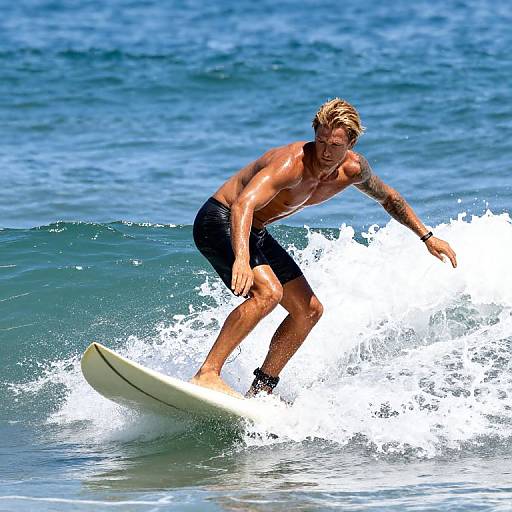 Photograph of a muscular, blonde, tattooed surfer in black board shorts riding a white surfboard on a blue ocean wave.