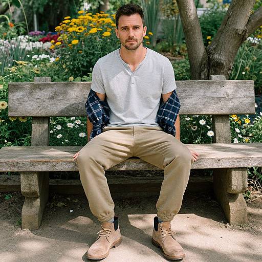 Photograph of a handsome, bearded man with short dark hair, wearing a white V-neck shirt, beige pants, and beige shoes, sitting on