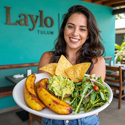 Smiling woman with long dark hair holds plate of grilled bananas, guacamole, and salad in turquoise restaurant with 