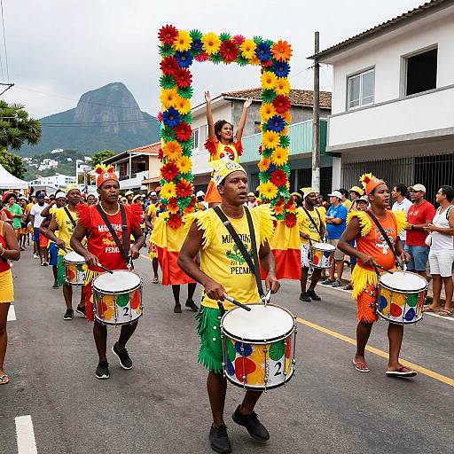 Colorful Carnival parade with drummers in yellow shirts, green skirts, and feathered headpieces, carrying a vibrant flower frame. Mountain in background.