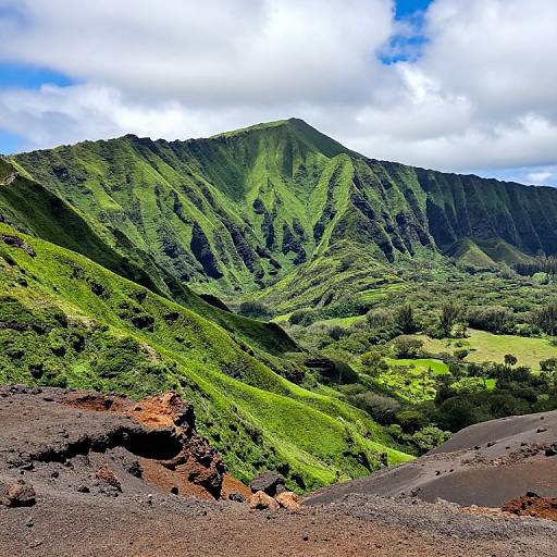 Photograph of vibrant green mountain with steep slopes, lush vegetation, and rocky foreground under a partly cloudy blue sky.
