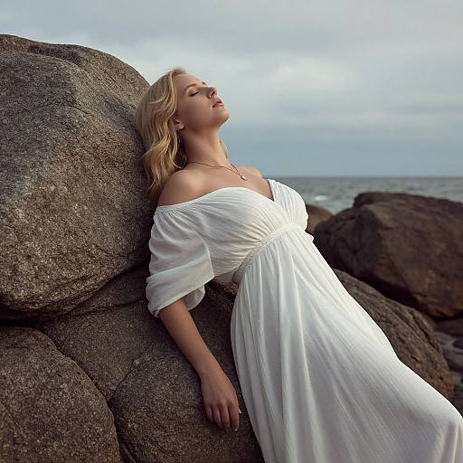 Blonde woman in white off-shoulder dress leans against large rocks by the sea, eyes closed, peaceful expression, cloudy sky background. Photographic