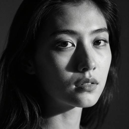 Black-and-white photograph of a young woman with dark hair, illuminated from the side, highlighting her serene expression and smooth skin. High contrast, dramatic lighting