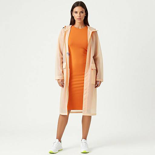Photograph of a straight-haired woman in an orange dress and beige trench coat, standing against a white background, wearing white sneakers.