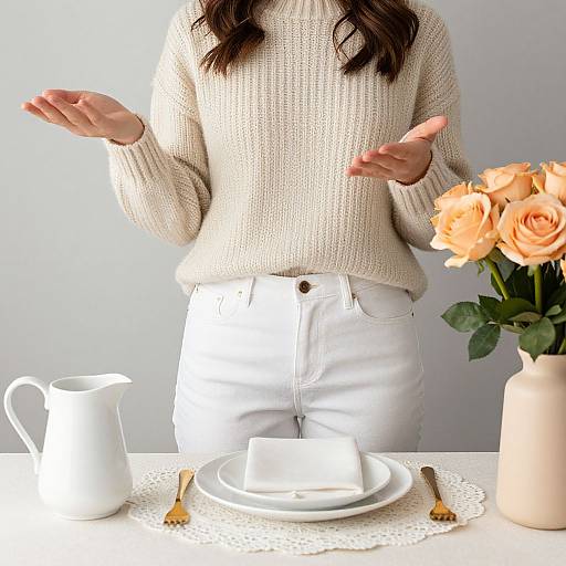 Photograph of a woman in a cream sweater and white pants, hands raised, standing by a table with a white teapot, plate, roses in