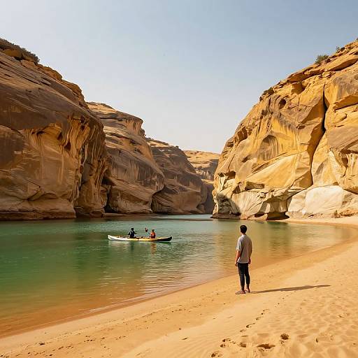 Photograph of a man standing on a sandy beach, facing a calm turquoise river, with a canoe and two people paddling between towering, sunlit