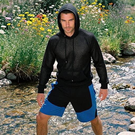 Photograph of a muscular man with a trimmed beard, wearing a black mesh hoodie and blue-black shorts, standing in a sunny, colorful flower-filled creek