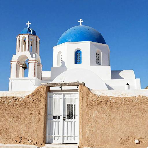 Photograph of a white Greek Orthodox church with blue domes and crosses, set against a clear blue sky, featuring a sandy-textured adobe wall