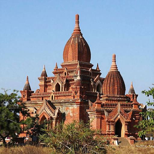 Photograph of a red brick Buddhist temple with multiple domes, intricate carvings, and arched windows, set against a clear blue sky,