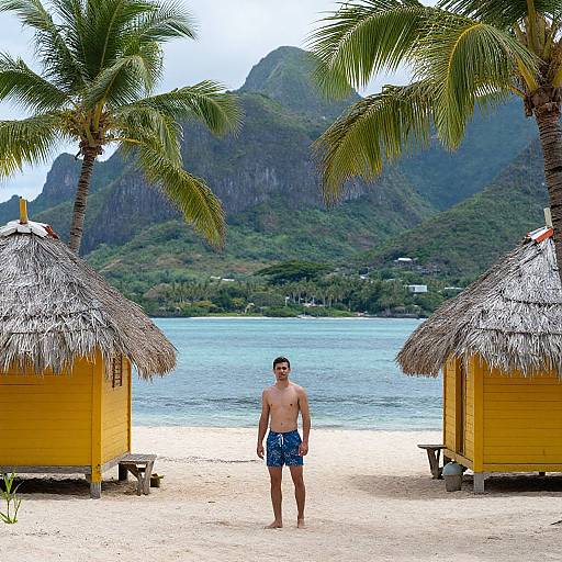 Photograph of a shirtless man in blue swim trunks standing on a tropical beach between two thatched-roof yellow huts, with palm trees