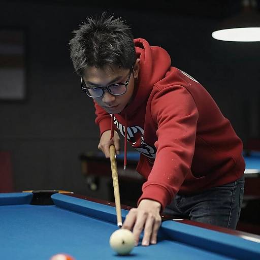 Photograph of a young Asian boy with black spiky hair, glasses, red hoodie, and jeans, aiming a pool cue at a ball on a