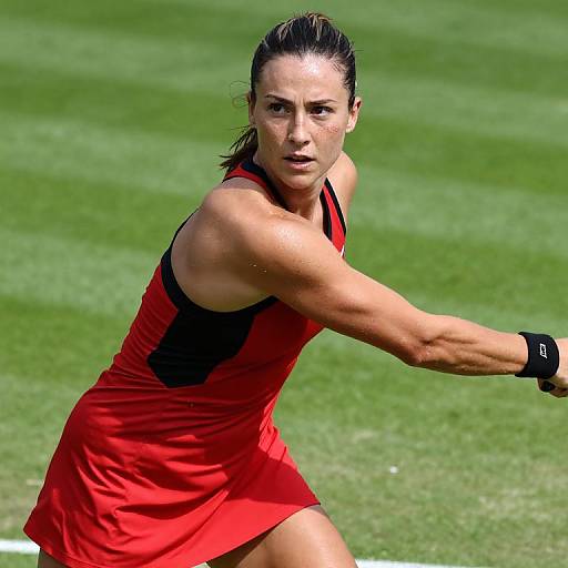 Photograph of a focused, muscular woman with wet brown hair, wearing a red sleeveless athletic dress, extending her arm to hit a tennis ball on