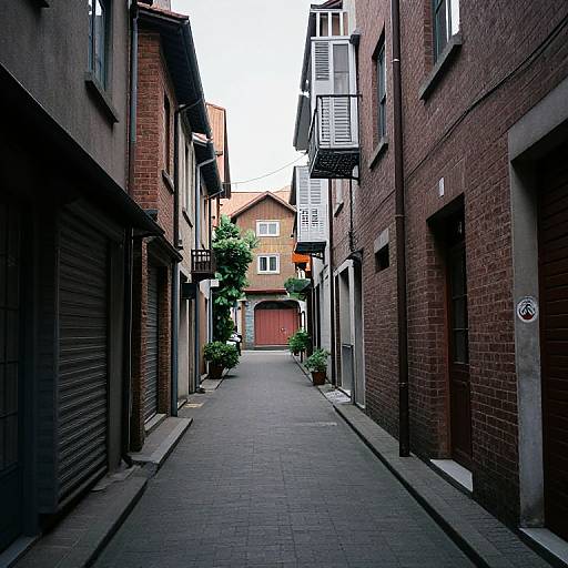 Photograph of a narrow, cobblestone alleyway between red-brick buildings, leading to a small, yellow house with a red garage door.
