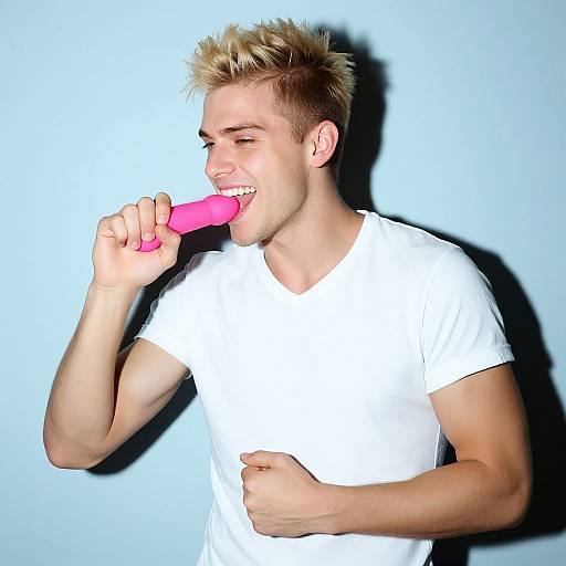 Photograph of a blond, muscular man in a white t-shirt, smiling while licking a pink vibrator against a bright blue background.