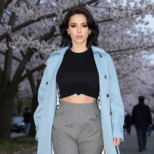 Photograph of a confident woman with dark hair, wearing a black crop top, light blue jacket, and high-waisted checkered pants, standing