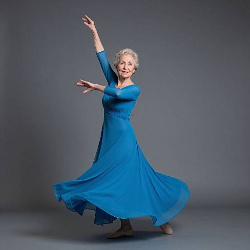 Elegant elderly woman with short gray hair, wearing a flowing blue dress, dancing gracefully against a gray studio backdrop.